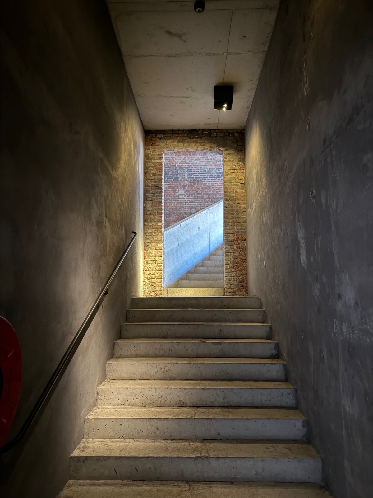 A narrow grey concrete staircase climbing towards a panel of old brick lit from a ceiling light.