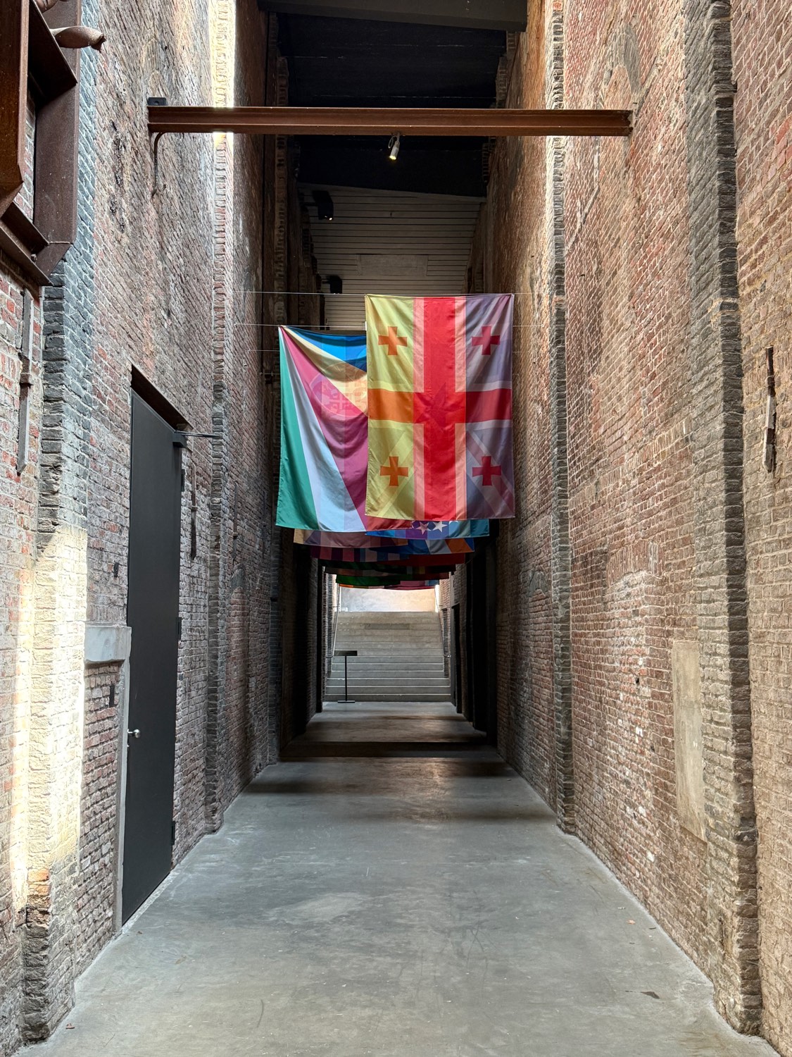 Fifteen patchwork fabric flags by Kimsooja hung in a line down a narrow brick corridor, receding towards light.