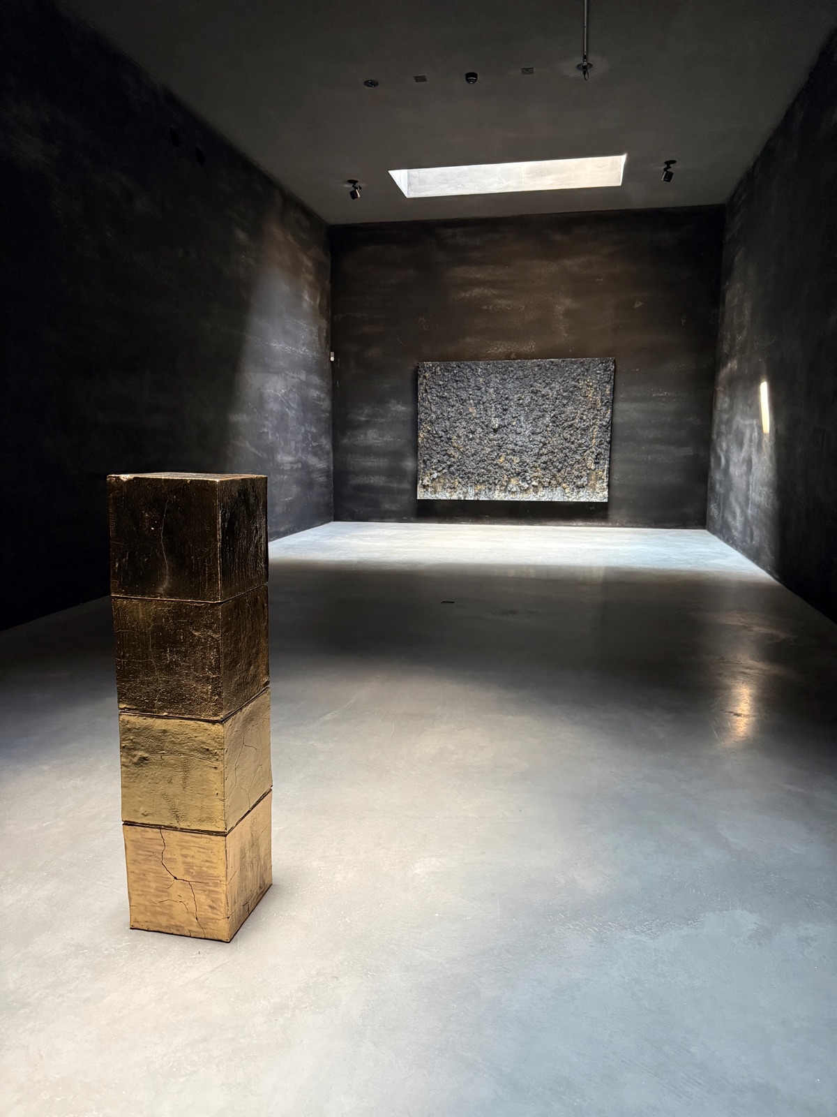 A gilded terracotta column by Bosco Sodi in a dark room, a thick cracked grey wall relief on the far wall.