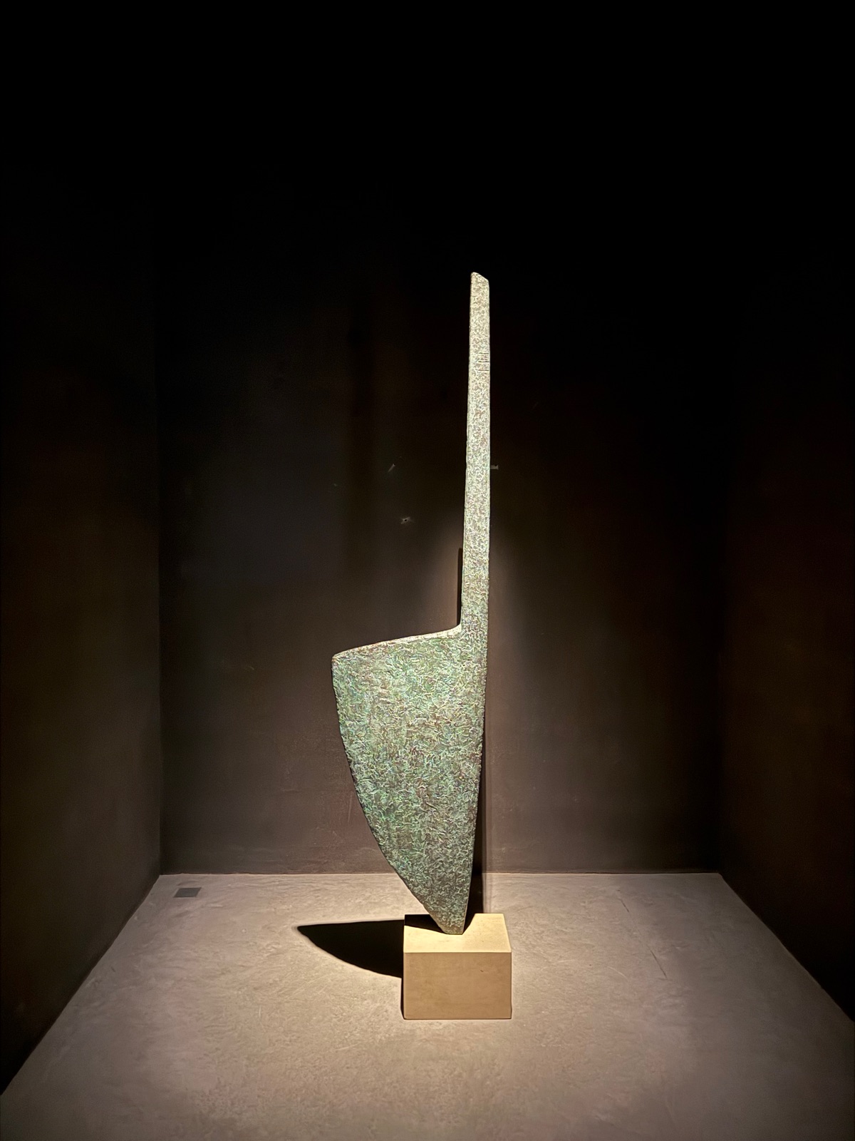 A tall blade-shaped bronze sculpture with green patina on a pale plinth, lit in a dark room.