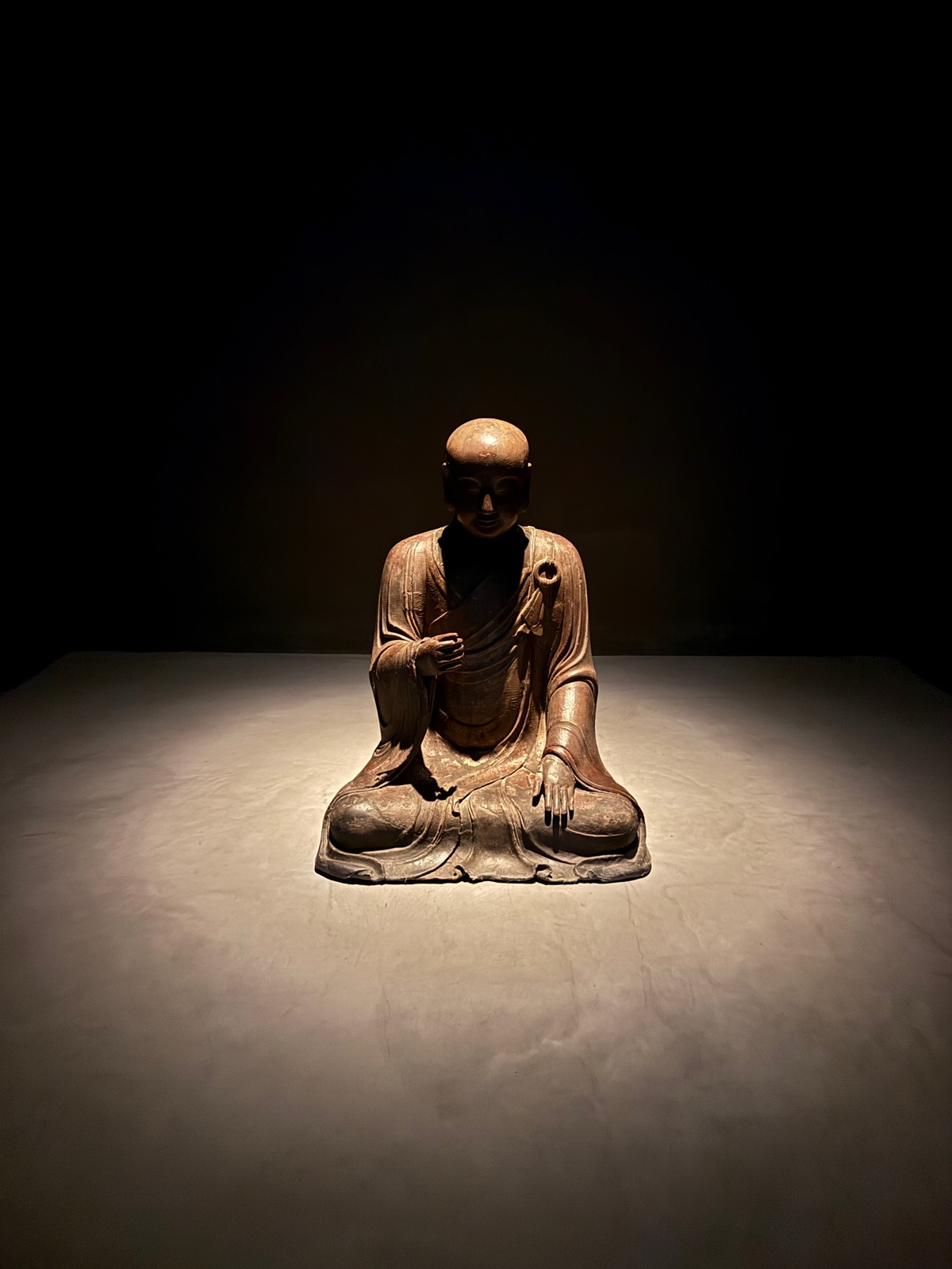 A small seated devotional figure, a bowed monk, carved in worn reddish wood, lit on a pale plinth in a dark room.