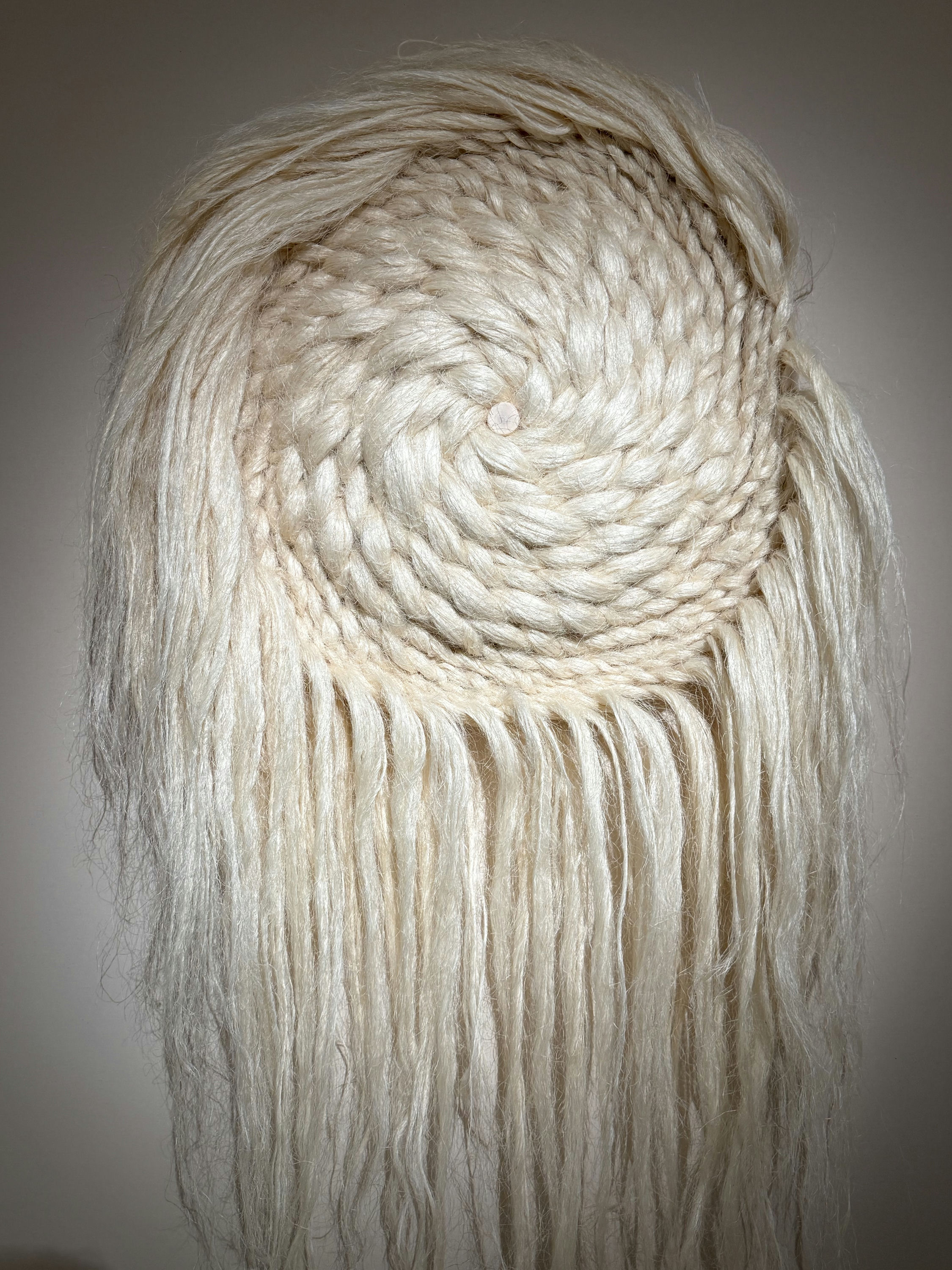 A spiralling cream textile work by Cécilie Bendixen, coiled tight at the centre and cascading into long loose fibres, on a pale wall.