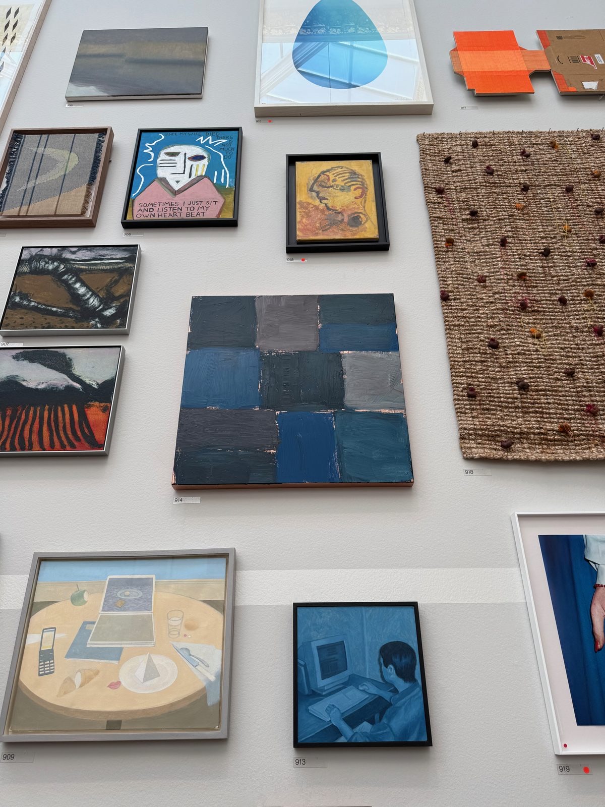 A small square abstract painting in cobalt blue and grey blocks, hung under a plain catalogue number among other works.