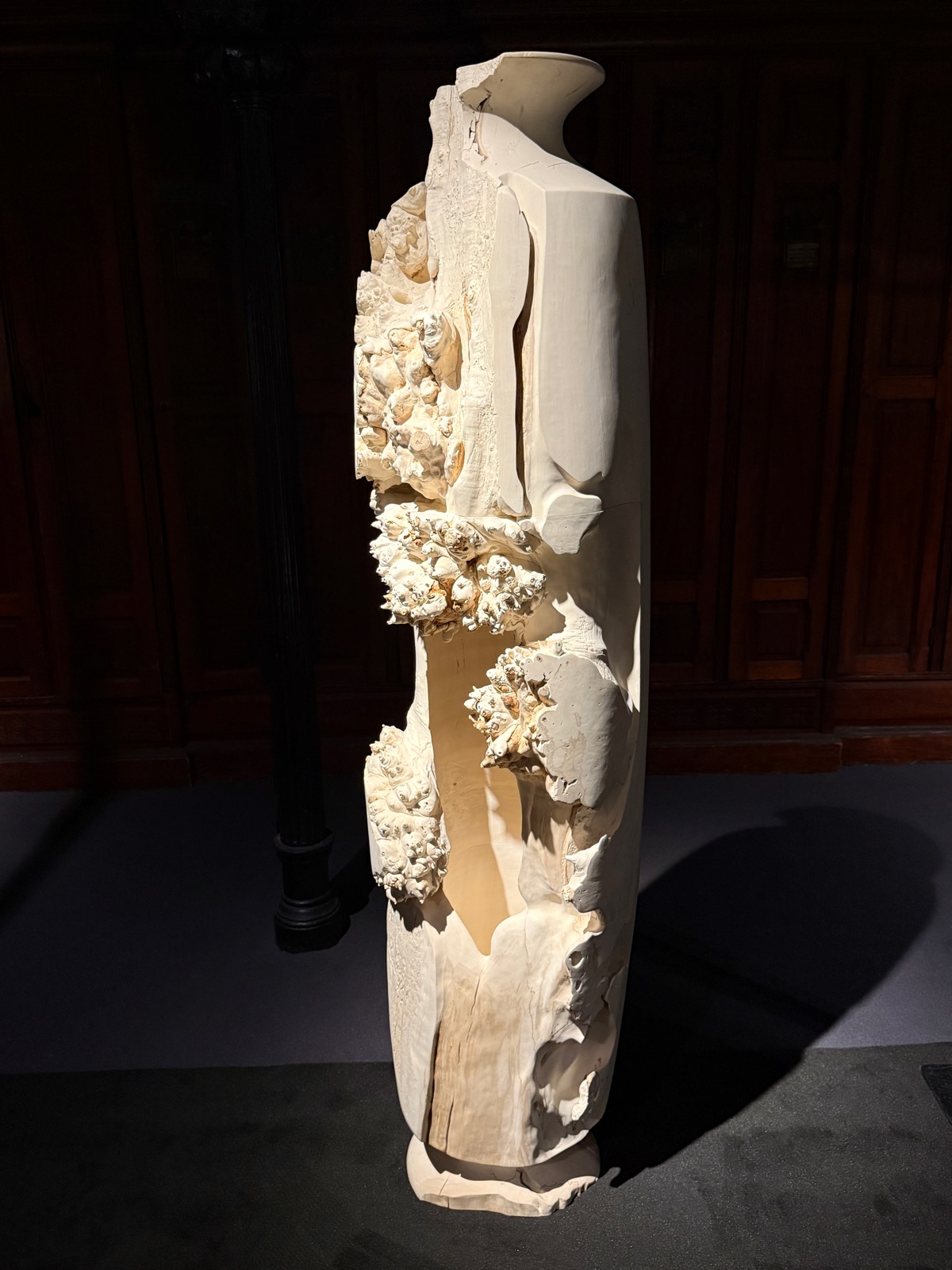 A tall pale bleached burr-wood column carved with barnacle-like clusters, lit against dark panelling.