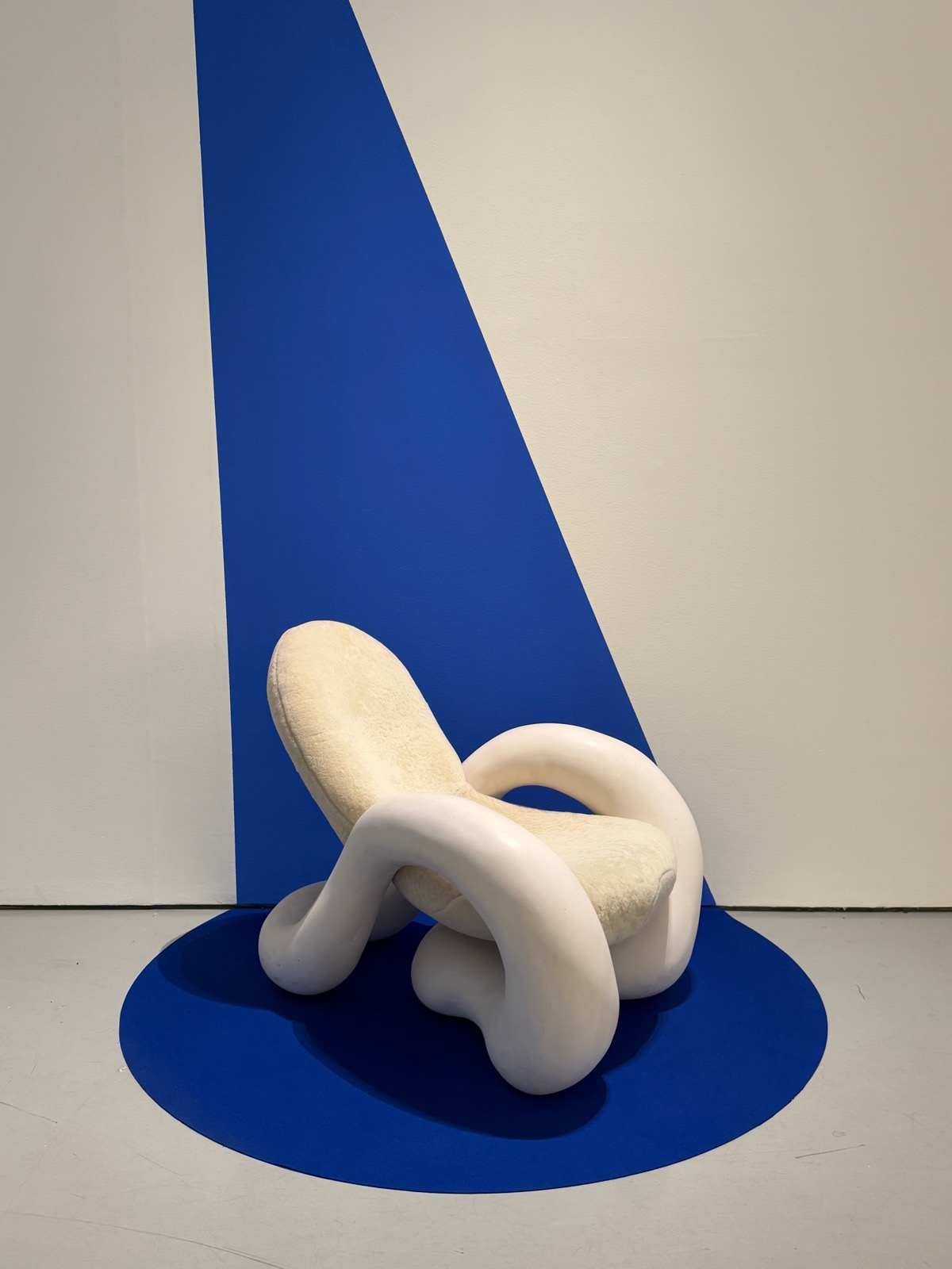 A smooth white looping sculptural chair with a fleece seat, lit on a cobalt-blue plinth.
