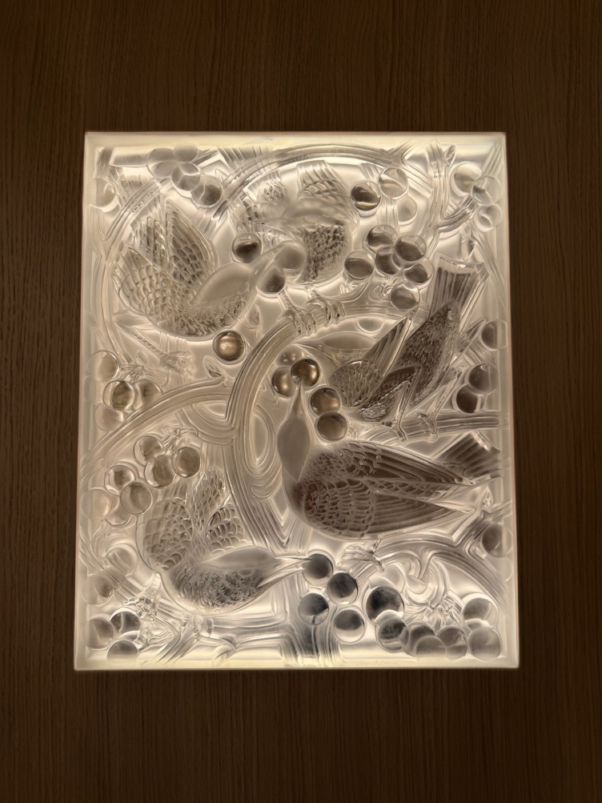 A backlit Lalique glass panel of blackbirds and grapes glowing behind the bar bottles.